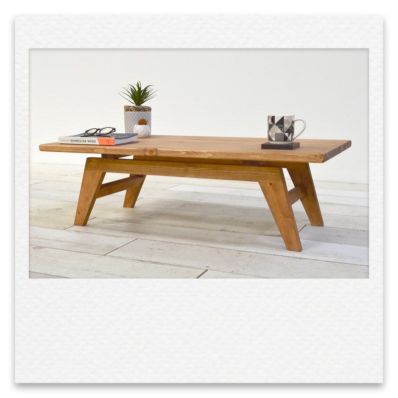 May include: A wooden coffee table with a natural wood finish and a rectangular top. The table has a simple design with four legs and a lower shelf. A book, a potted plant, and a mug are on the table.