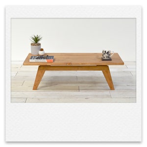May include: A light brown wooden coffee table with a rectangular top and a lower shelf. The table has four legs and is sitting on a light wood floor. There is a book, a potted plant, and a mug on the table.