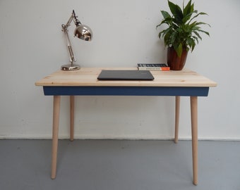 May include: A wooden desk with a blue painted bottom edge and natural wood top. The desk has four light wood legs and a laptop computer on the surface. A lamp and a book are also on the desk.