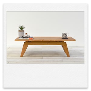 May include: A wooden coffee table with a natural finish and a simple design. The table has a rectangular top and four legs. There are a few items on the table, including a book, a pair of glasses, and a mug.