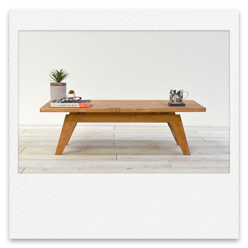 May include: A wooden coffee table with a natural finish and a simple design. The table has a rectangular top and four legs. There are a few items on the table, including a book, a pair of glasses, and a mug.