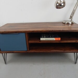 May include: A dark brown wooden coffee table with a blue painted drawer and two shelves. The table has hairpin legs and a book titled "Norwegian Wood" on the top shelf.