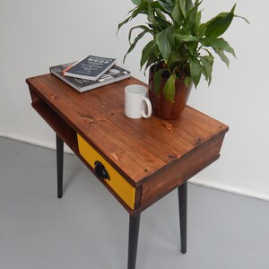 May include: A small wooden table with a yellow drawer and black legs. The table has a book on top and a white mug. A green plant in a brown pot is on the table.