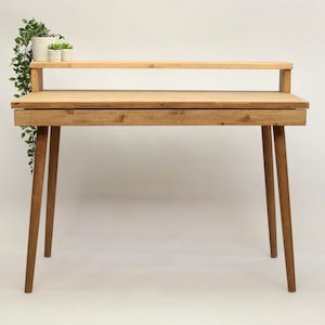 May include: A light wood desk with a shelf above the desktop. The desk has four legs and a drawer. There are two small potted plants on the shelf.