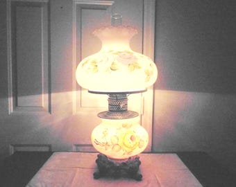 May include: A vintage oil lamp with a white glass shade, adorned with hand-painted floral designs in pink and yellow. The lamp has a decorative metal base and a ruffled top. The lamp is lit, casting a warm glow.