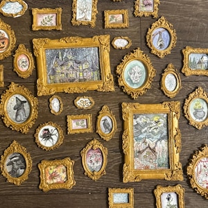 Puede incluir: Una colección de pequeñas obras de arte enmarcadas en marcos dorados ornamentados. Las ilustraciones presentan temas góticos y espeluznantes, como una casa encantada, murciélagos y calaveras. Los marcos están dispuestos sobre una superficie de madera.