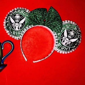 Op de afbeelding: Een groen en wit Tinkerbell-hoofdband met een wit hoofdband en groene glitteroren. De hoofdband heeft de tekst "2nd Star to the Right" en een Tinkerbell-afbeelding op elk oor. Een groene en witte theekop met een sneeuwvlokkenontwerp is ook op de afbeelding te zien.