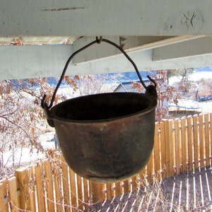 May include: A rusty metal pot with a wire handle hanging from a wooden beam. The pot is empty and has a dark brown color. The pot is hanging in front of a wooden fence and a blurry background of trees and a house.