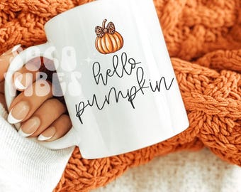 May include: White ceramic mug with a pumpkin illustration and the words "hello pumpkin" in black script. The mug has a handle and is set against an orange knitted blanket. The pumpkin has a leopard print bow.