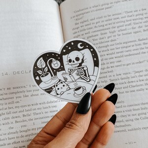 May include: A heart-shaped sticker with a black and white illustration of a skeleton reading a book, with a cat, plant, and coffee cup. The sticker is held over an open book with text.