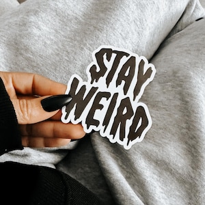 May include: A die-cut sticker featuring the text STAY WEIRD in a bold, black, dripping font style with a white border outline. The lettering has a distinct liquid or melting appearance, creating a high-contrast graphic design on a white background.