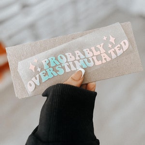 May include: A sticker with the text "Probably Overstimulated" in a rainbow gradient of pink, blue, and green. The sticker is on a brown paper background.