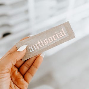 May include: A brown paper sticker with the word "antisocial" printed in pink gothic lettering.