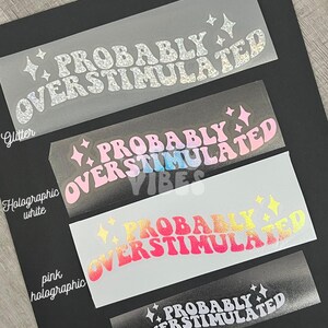May include: Four clear vinyl stickers with the text "Probably Overstimulated" in a retro font. The stickers are in different colors: glitter, holographic white, pink holographic, and white.
