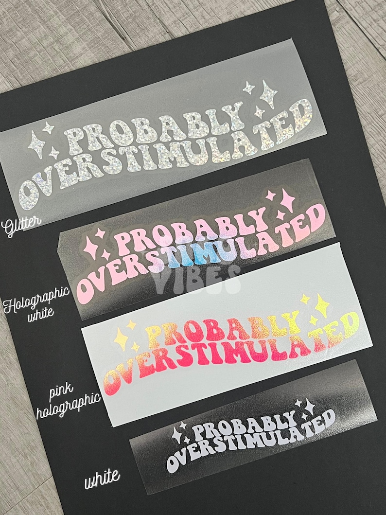 May include: Four clear vinyl stickers with the text "Probably Overstimulated" in a retro font. The stickers are in different colors: glitter, holographic white, pink holographic, and white.