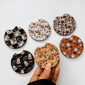 May include: A collection of six round car coasters with Halloween-themed designs. The coasters feature patterns of ghosts, pumpkins, bats, and spiderwebs in various colors, including black, orange, and white. One coaster is held by a person. These coasters are designed to fit in a car's cup holder.