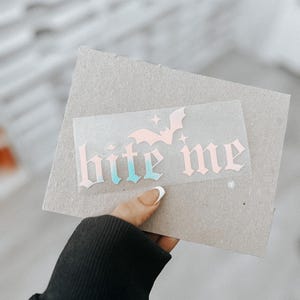 May include: A pink and blue vinyl sticker with the text "bite me" in a gothic font. A bat silhouette is above the text.