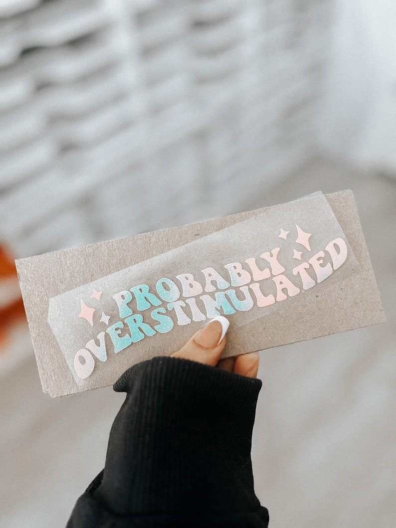 May include: A sticker with the text "Probably Overstimulated" in a rainbow gradient of pink, blue, and green. The sticker is on a brown paper background.