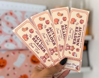 May include: Four beige "Autumn Reader Book Club" bookmarks with fall-themed illustrations of pumpkins, socks, and teddy bears. The text "Admit One" and "cozy girlie" are also visible.