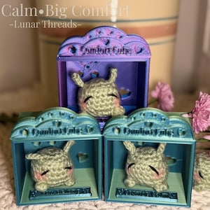May include: Three Comfort Cube displays with crocheted creatures. The cubes are blue and purple, with the text "Comfort Cube" on top. The creatures are gray with pink cheeks and black eyes. The image also includes pink flowers and the text "Little Calm • Big Comfort - Lunar Threads."