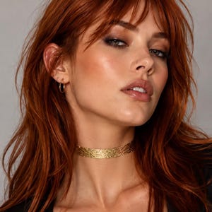 May include: A gold choker necklace featuring a textured, shimmering surface that wraps closely around the neck. The piece has a metallic finish with a fine, intricate pattern that catches the light, creating a subtle sparkle against the skin.