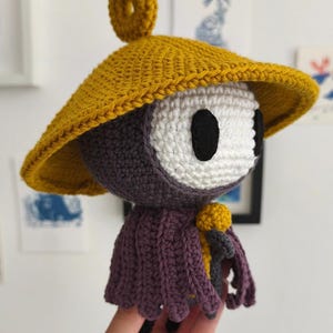 May include: Crocheted doll with a yellow hat and a purple cloak. The doll has a white face with black eyes and a yellow ring on top of the hat. The doll is handmade and has a unique design.
