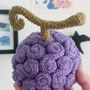 Könnte beinhalten: Eine gehäkelte lila Frucht mit strukturierter Oberfläche und braunem Stiel. Das handgefertigte Objekt wird in einer Hand gehalten. Die Oberfläche der Frucht ist mit spiralförmigen Häkelstichen bedeckt. Der Stiel ist hellbraun.