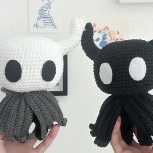 May include: Two handmade crochet plush toys, one white and one black, each with large oval eyes and horn-like appendages. The white toy has a grey cape, while the black one has a similar cape. These unique, handcrafted items are perfect for fans of video games.
