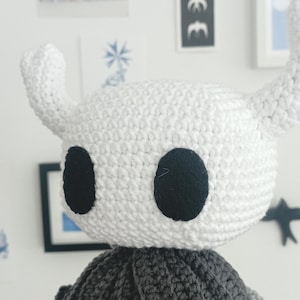 Puede incluir: Muñeca de ganchillo hecha a mano con una cabeza blanca, dos cuernos curvos y ojos negros grandes. La muñeca tiene una parte inferior gris y con flecos. El fondo incluye obras de arte enmarcadas y elementos decorativos.