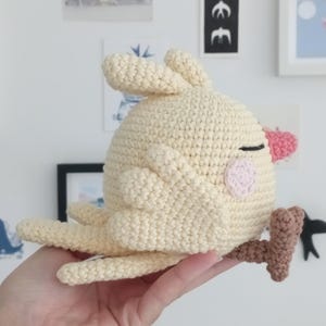 May include: A crocheted yellow bird with pink cheeks and a brown twig in its beak. The bird is sitting on a hand.
