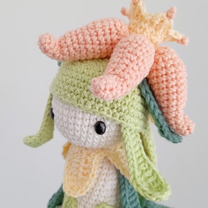 May include: A crocheted plush toy with a green and white body, a pink flower on top of its head, and a yellow flower on its chest.