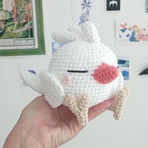 May include: White crocheted bird plush toy with pink cheeks and a pink beak. The bird has a black eye and is sitting on a hand.