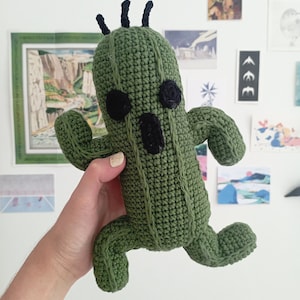 May include: A green crocheted cactus plush toy with black eyes and three black spikes on top of its head. The cactus is standing on its two arms and legs.
