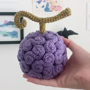 May include: A handmade, crocheted purple fruit with a stem and curly top. The fruit is a deep lavender color, with a textured surface created by the crochet stitches. The stem and top are a light brown color.