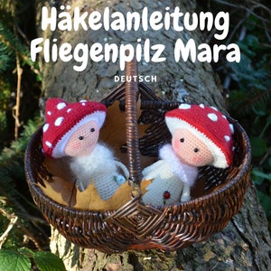 Könnte beinhalten: Zwei gehäkelte Pilzpuppen mit weißen Gesichtern und roten und weißen Pilzhüten sitzen in einem braunen Weidenkorb. Der Korb steht auf einem Baumstamm. Der Text "Häkelanleitung Fliegenpilz Mara DEUTSCH" ist über dem Korb zu sehen.