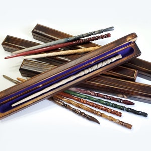 May include: A collection of slender, carved wooden wands in various colors including red, white, green, brown, and black, each featuring unique textured handles. One light-colored wand rests inside an open, rectangular wooden box lined with purple velvet.