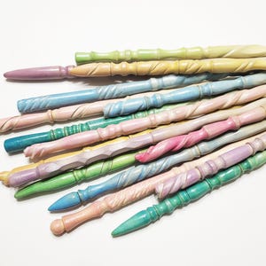 Pode incluir: Uma cole&ccedil;&atilde;o de varinhas de madeira em cores pastel, em tons de rosa, azul, verde e amarelo. Cada varinha tem um design esculpido &uacute;nico, com padr&otilde;es torcidos e texturizados. As varinhas est&atilde;o dispostas sobre uma superf&iacute;cie branca.