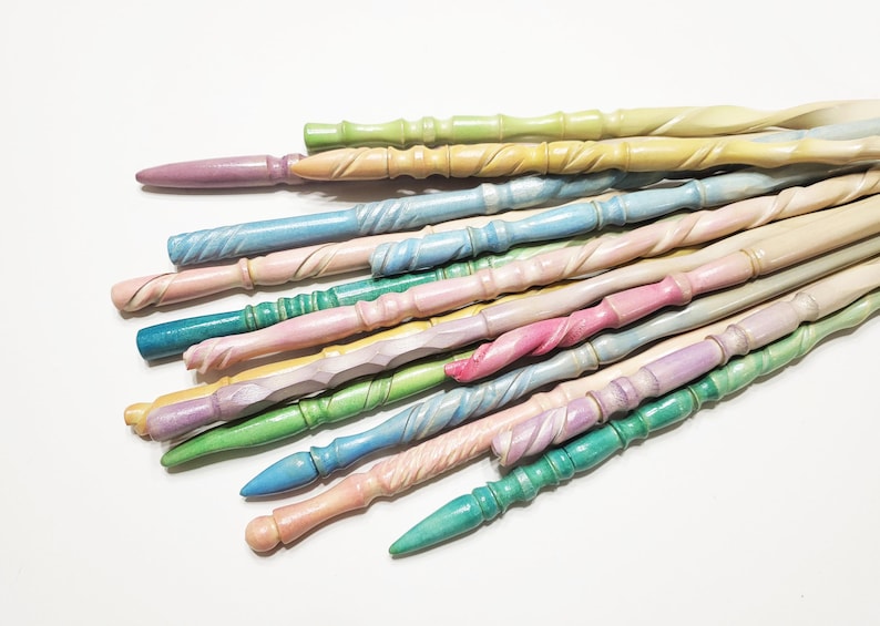 Pode incluir: Uma cole&ccedil;&atilde;o de varinhas de madeira em cores pastel, em tons de rosa, azul, verde e amarelo. Cada varinha tem um design esculpido &uacute;nico, com padr&otilde;es torcidos e texturizados. As varinhas est&atilde;o dispostas sobre uma superf&iacute;cie branca.