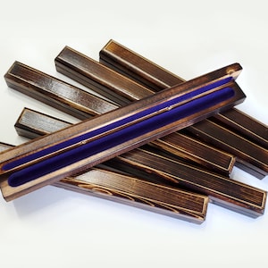 May include: A collection of seven long, rectangular wooden boxes with a dark brown, stained finish and visible wood grain. Each box features a hinged lid and a deep purple velvet-lined interior channel designed to hold a single slender object.
