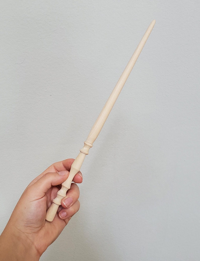 May include: A plain wooden wand with a rounded tip and a detailed handle. The wand is held in a hand.