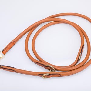 May include: A tan leather dog leash with a rounded, tubular design. The leash features gold-toned metal hardware, including a swivel snap hook at one end and two adjustable metal rings along the length of the strap for versatile use.