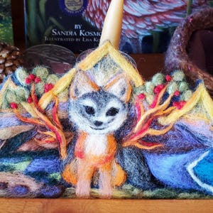 May include: A felt crown with a fox in the center, surrounded by a landscape with trees and mountains. The fox is orange and white with black eyes. The crown is yellow, orange, blue, and green.