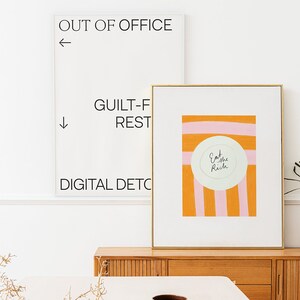 Op de afbeelding: Een ingelijst print met een roze en oranje gestreepte achtergrond en een witte cirkel met de tekst "Eat the Rich" in zwarte cursieve letters. De print hangt aan een muur boven een houten dressoir.