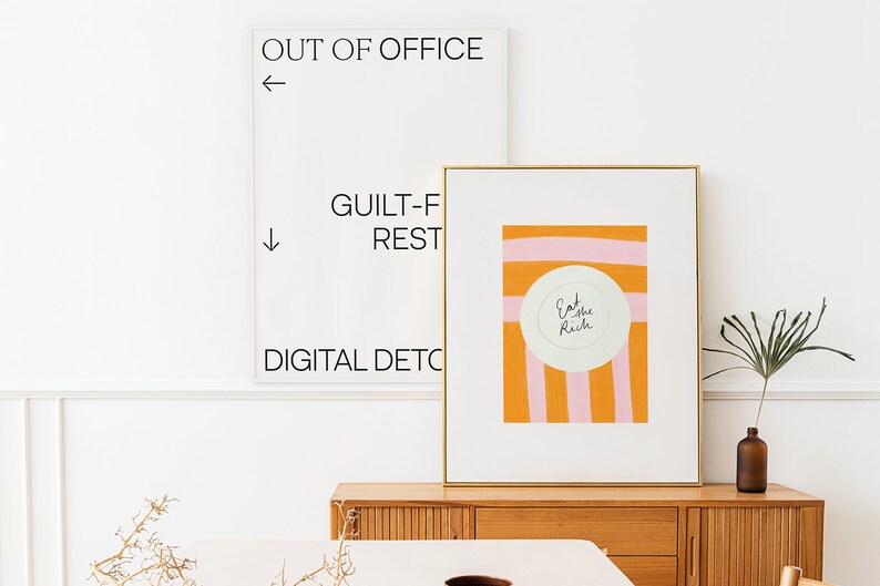 Op de afbeelding: Een ingelijst print met een roze en oranje gestreepte achtergrond en een witte cirkel met de tekst "Eat the Rich" in zwarte cursieve letters. De print hangt aan een muur boven een houten dressoir.