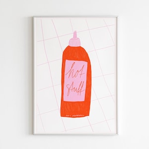 May include: A framed wall art print featuring a bright orange squeeze bottle with a pink cap and a pink rectangular label. The label displays the handwritten text hot stuff in red cursive. The background is white with a thin pink grid pattern.
