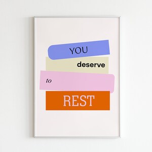 May include: A white framed poster with the text "YOU deserve to REST" in black and white text on a stack of colourful rectangles. The rectangles are blue, beige, pink, and orange.