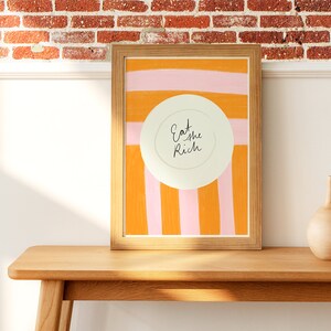 Op de afbeelding: Een ingelijst print met een witte cirkel in het midden waarop "Eat the Rich" staat, op een achtergrond van oranje en roze strepen.