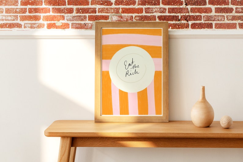Op de afbeelding: Een ingelijst print met een witte cirkel in het midden waarop "Eat the Rich" staat, op een achtergrond van oranje en roze strepen.