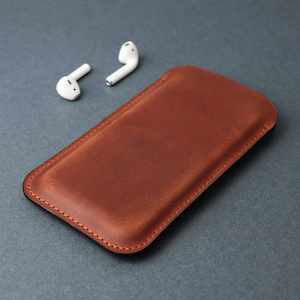 May include: A brown leather phone case with a stitched edge. The case is lying on a gray surface next to two white wireless earbuds.
