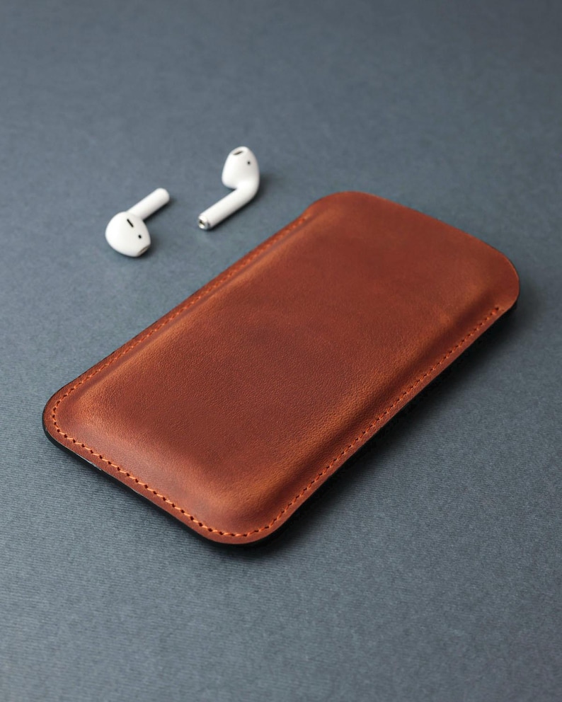 May include: A brown leather phone case with a stitched edge. The case is lying on a gray surface next to two white wireless earbuds.